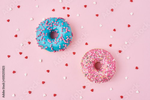 Two donuts decorated icing and sprinkles and confetti in the shape of heart on pink background