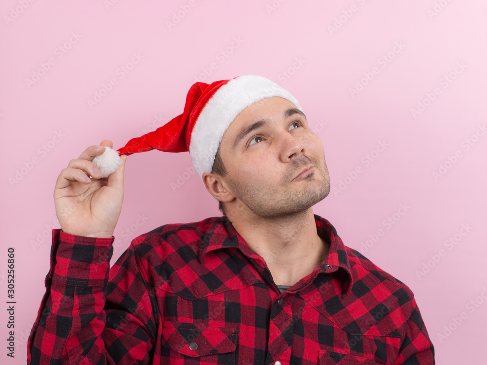 Emotions on the face, pensive, reflection, plan, idea. A man in a plaid rabbit and a Christmas red hat, on a pink background, copy space. Emotions on the face, pensive, reflection, plan, idea. A man in a plaid rabbit and a Christmas red hat, on a pink background, copy space.