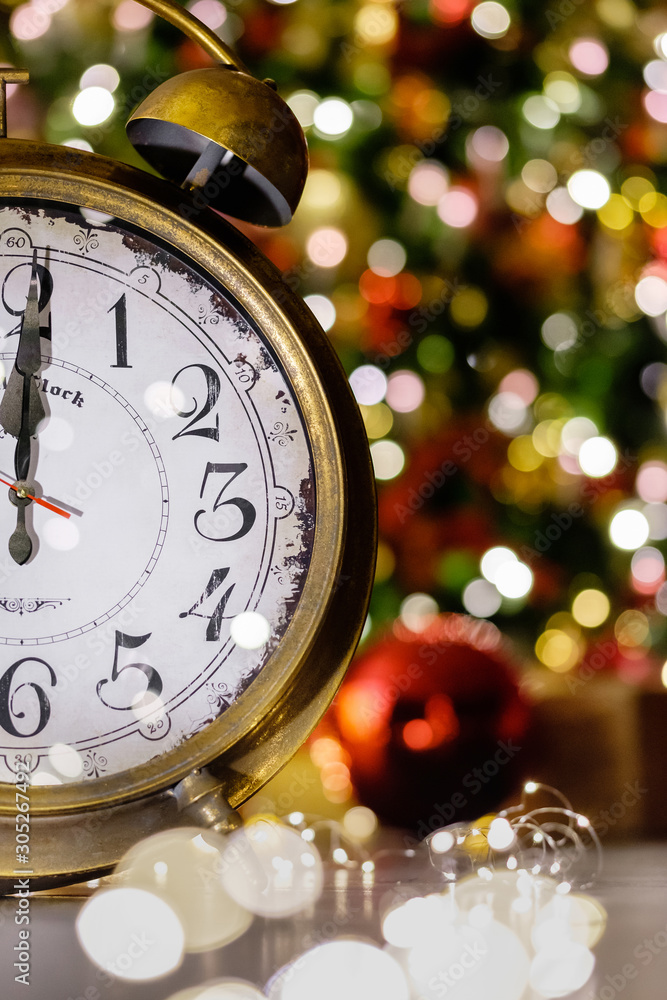 Close-up fragment of golden clock showing twelve o'clock standing on floor next to burning golden lights on backdrop of New Year's decor and Christmas tree. Concept of expectation of a miracle