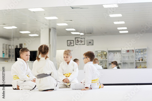 Group of children in kimono sitting on the floor and talking to each other during their lesson in karate in the gym