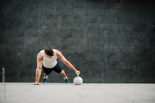 Dedicated handsome muscular caucasian man in shorts and t-shirt doing push ups with kettle bell. In background is gray wall.