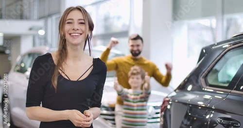 Young beautiful Caucasian woman bragging car keys and smiling. Her husband and son showing thumbs up at the background. Successful family buying new vehicle in showroom. Cinema 4k footage ProRes HQ.