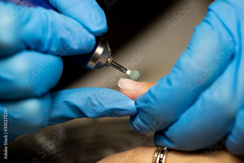 Hands in blue rubber gloves are holding a tool for a hardware manicure over a long nail close-up.