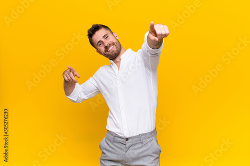 young handosme man feeling happy and confident, pointing to camera with both hands and laughing, choosing you against flat color wall