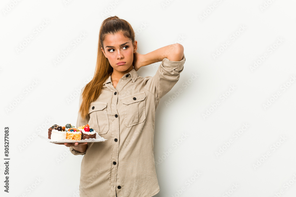 Young caucasian woman holding sweet cakes touching back of head, thinking and making a choice. Young caucasian woman holding sweet cakes touching back of head, thinking and making a choice.