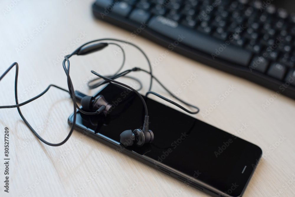 Black smartphone and headphone in the foreground, against a blurred background with a computer keyboard. Black smartphone and headphone in the foreground, against a blurred background with a computer keyboard.
