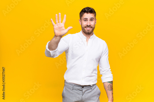 young handosme man smiling and looking friendly, showing number five or fifth with hand forward, counting down against flat color wall