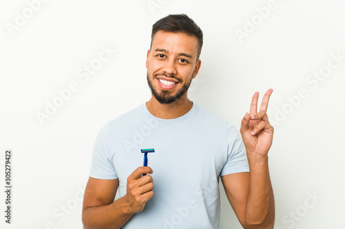 Young south-asian man holding a razor blade joyful and carefree showing a peace symbol with fingers.
