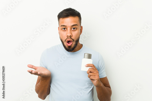 Young south-asian man holding an after shave cream surprised and shocked.