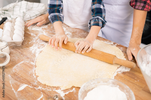 Hands of child with wooden pin rolling fresh dough on table while helping mom