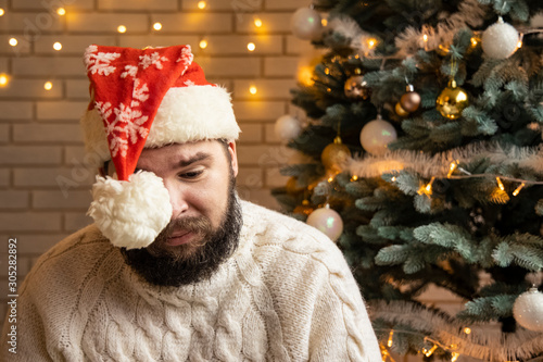 Alone on Christmas concept depression and stressful sad emotions by bearded young adult man hipster style in Santa Claus in room with garland lights background  photo