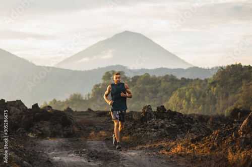 Young athlete man trail running in mountains in the morning. Amazing volcanic landscape of Bali mount Agung on background. Healthy lifestyle concept.