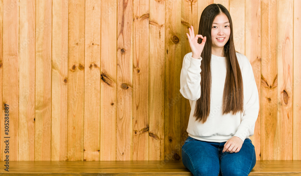 Young chinese woman sitting on a wooden place cheerful and confident showing ok gesture.