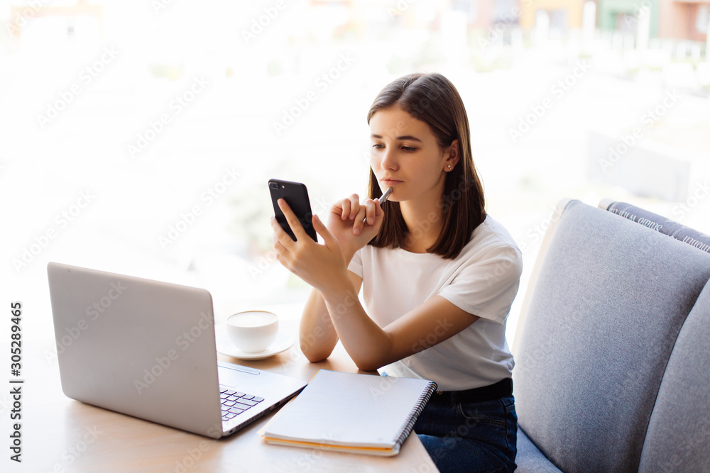 Young pretty woman sitting in coffee shop with laptop at wooden table, drinking coffee and using smartphone. Young pretty woman sitting in coffee shop with laptop at wooden table, drinking coffee and using smartphone.