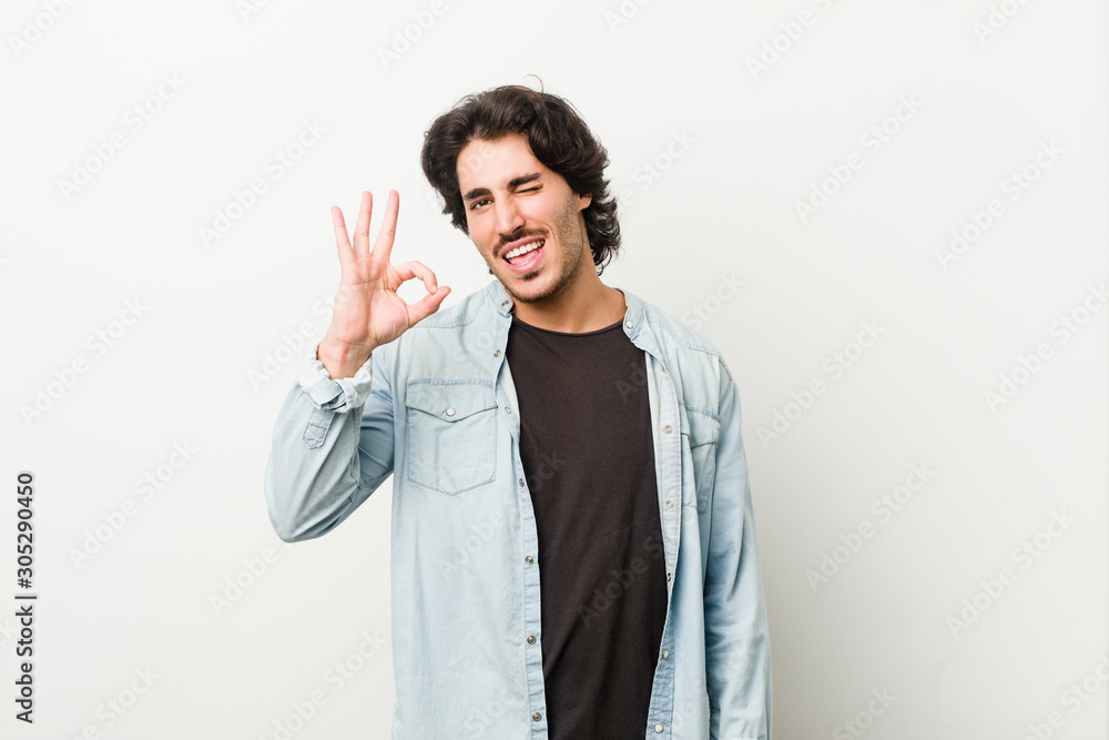 Young handsome man against a white background winks an eye and holds an okay gesture with hand. Young handsome man against a white background winks an eye and holds an okay gesture with hand.