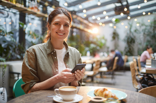 Portrait of beautiful young woman smiling at camera while using smartphone on outdoor terrace in cafe or coffee shop, copy space