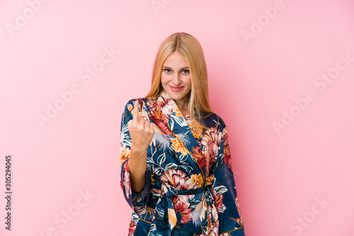 Young blonde woman wearing a kimono pajama pointing with finger at you as if inviting come closer.