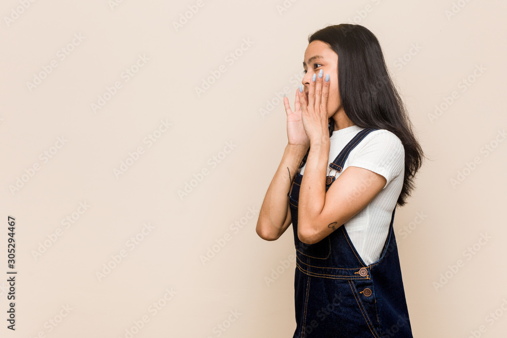 Young cute chinese teenager Young blonde woman wearing a coat against a pink background shouts loud, keeps eyes opened and hands tense.