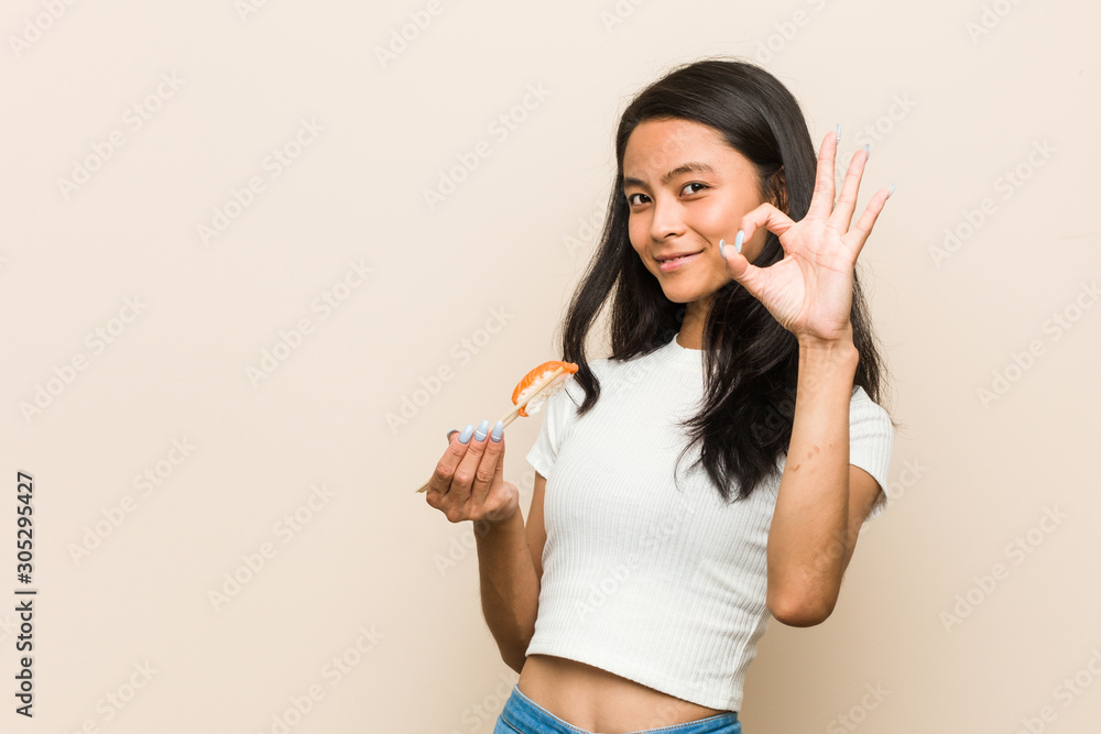Young asian woman holding a sushi piece cheerful and confident showing ok gesture. Young asian woman holding a sushi piece cheerful and confident showing ok gesture.