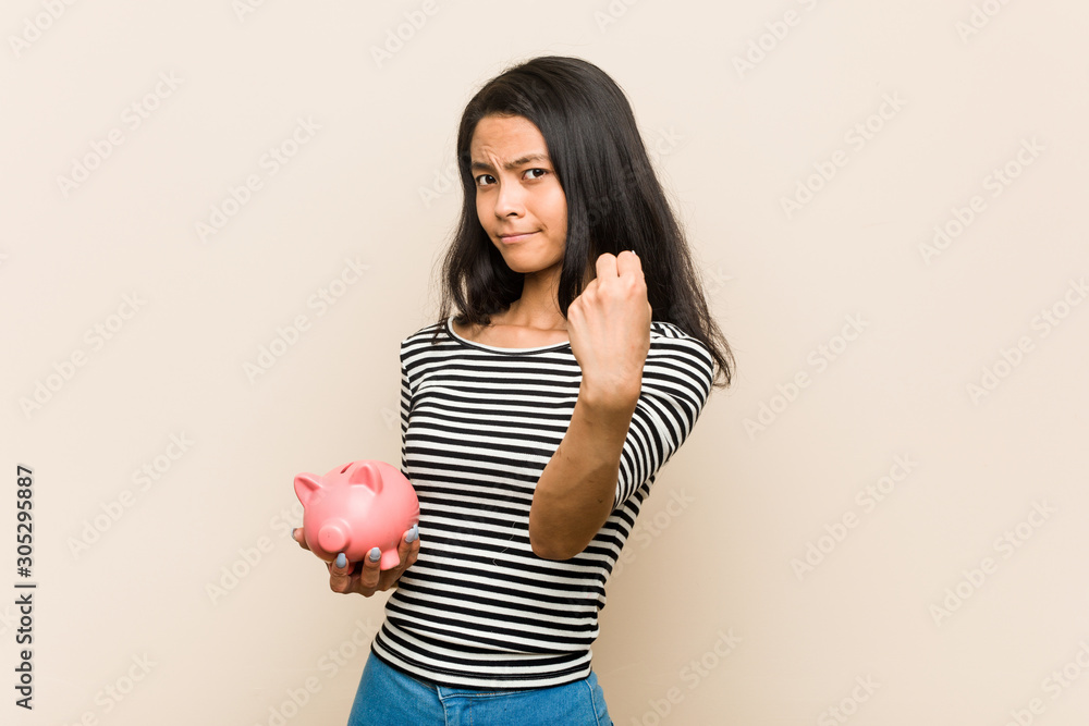 Young asian woman holding a piggy bank showing fist to camera, aggressive facial expression. Young asian woman holding a piggy bank showing fist to camera, aggressive facial expression.
