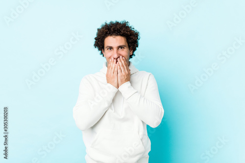 Young sporty man against a blue background laughing about something, covering mouth with hands.