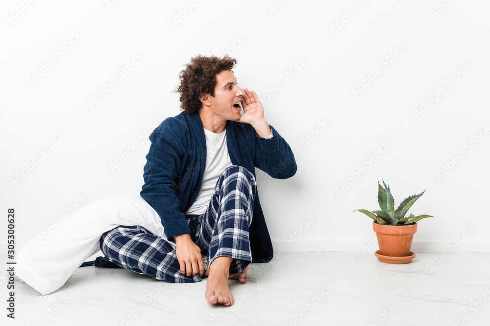 Mature man wearing pajama sitting on house floor shouting and holding palm near opened mouth.