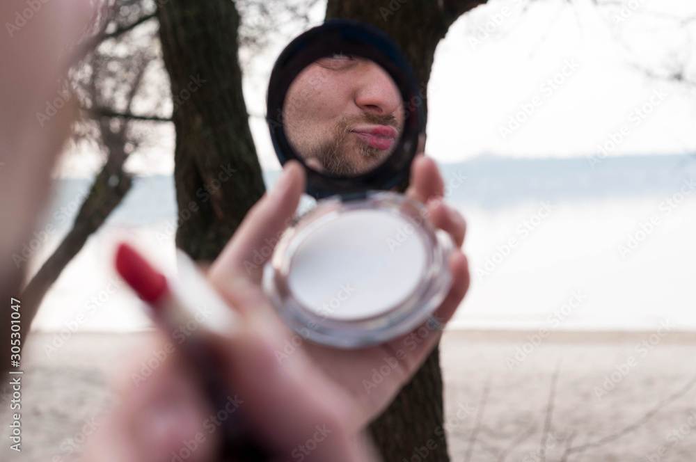 soft focus of man reflection in pocket mirror applying red lipstick in front of trees and river soft focus of man reflection in pocket mirror applying red lipstick in front of trees and river