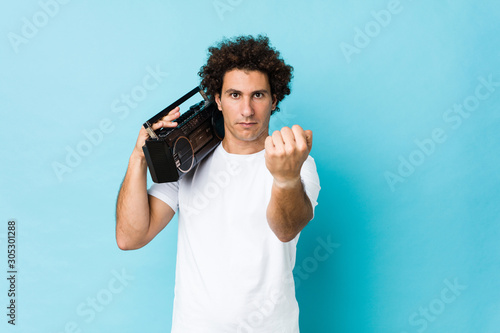 Young caucasian curly man holding a guetto blaster showing fist to camera, aggressive facial expression.