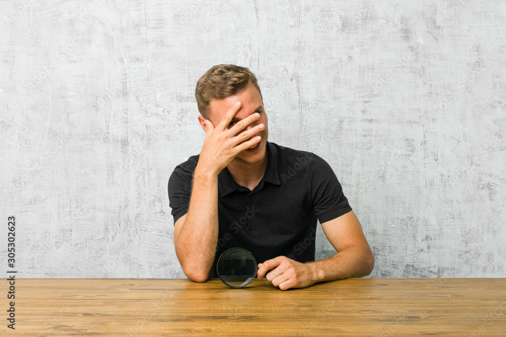 Young handsome man holding a magnifying glass blink at the camera through fingers, embarrassed covering face. Young handsome man holding a magnifying glass blink at the camera through fingers, embarrassed covering face.