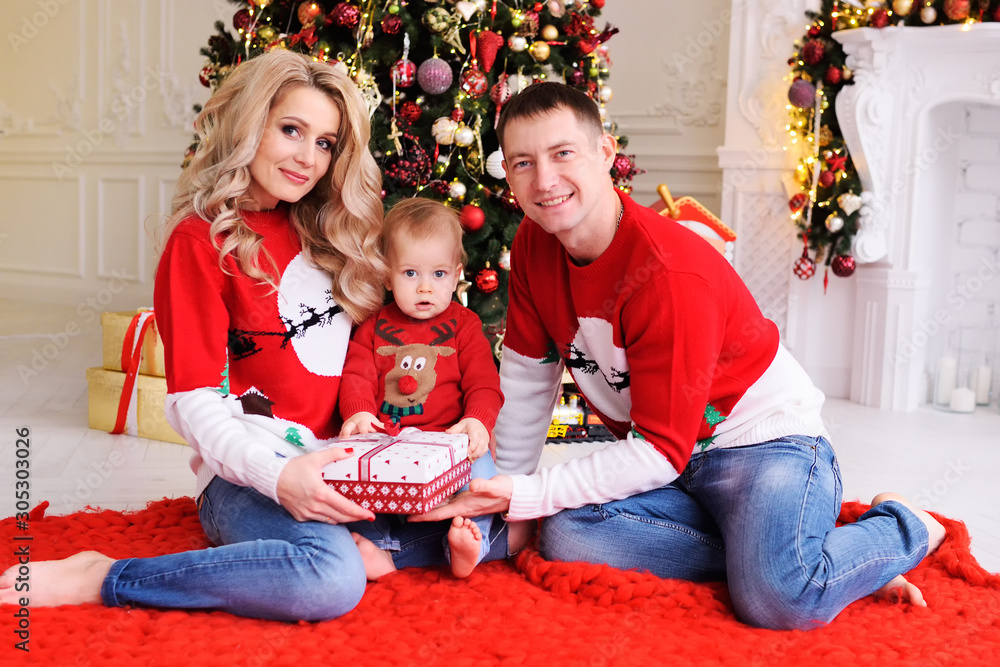 young family-mom, dad and little son in Christmas sweaters with reindeer on the background of the Christmas tree. Parents give a Christmas gift to their child. young family-mom, dad and little son in Christmas sweaters with reindeer on the background of the Christmas tree. Parents give a Christmas gift to their child.