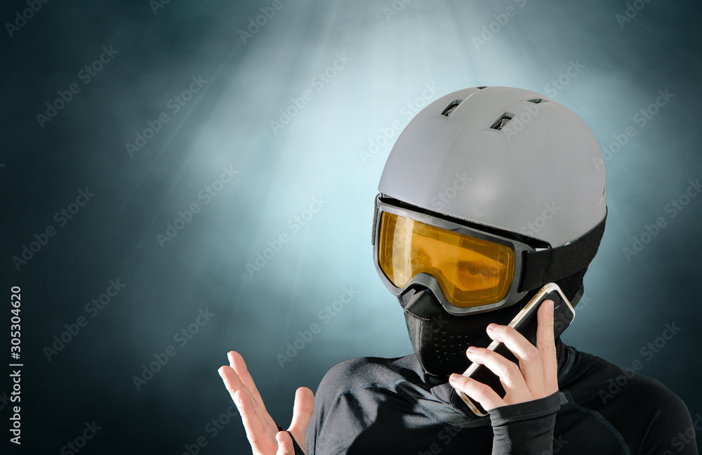 Athlete wearing a sweatshirt, mask and helmet talking on the phone on a dark background. The concept of conversation, determining the details. Practicing dangerous sports, proper clothing.