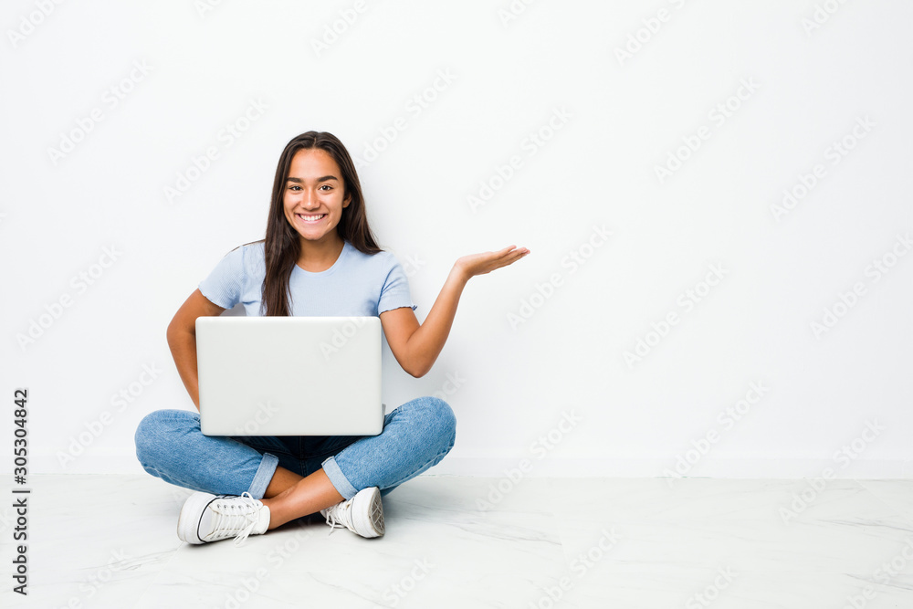 Young mixed race indian woman sitting working on laptop showing a copy space on a palm and holding another hand on waist. Young mixed race indian woman sitting working on laptop showing a copy space on a palm and holding another hand on waist.