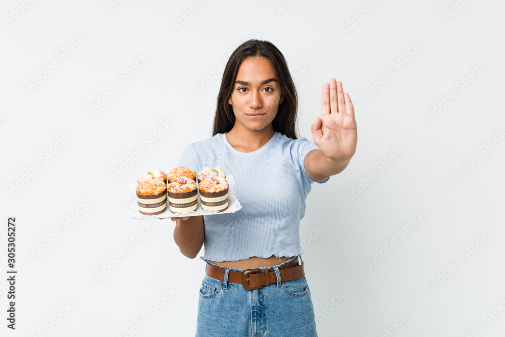 Young mixed race indian holding a sweet cakes standing with outstretched hand showing stop sign, preventing you.