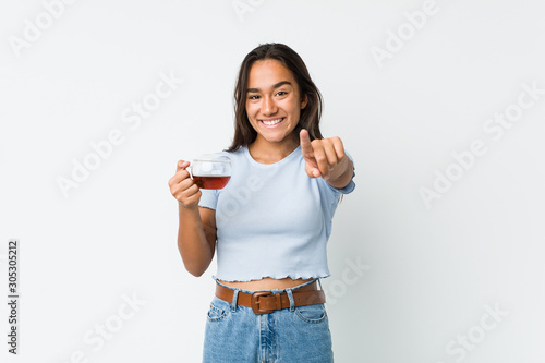 Young mixed race indian holding a tea cup cheerful smiles pointing to front.