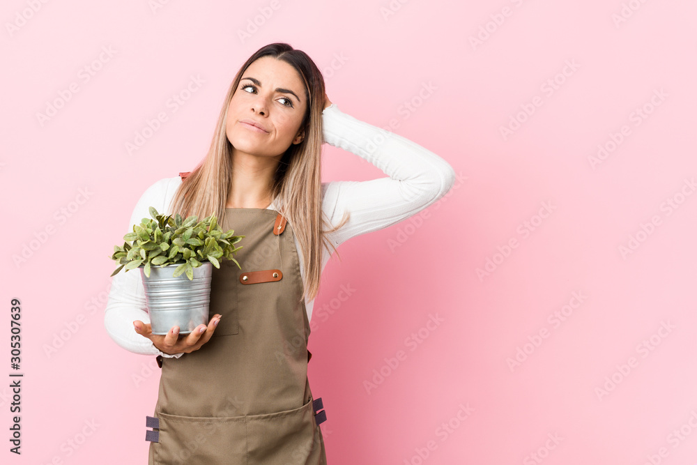 Young gardener woman holding a plant touching back of head, thinking and making a choice. Young gardener woman holding a plant touching back of head, thinking and making a choice.