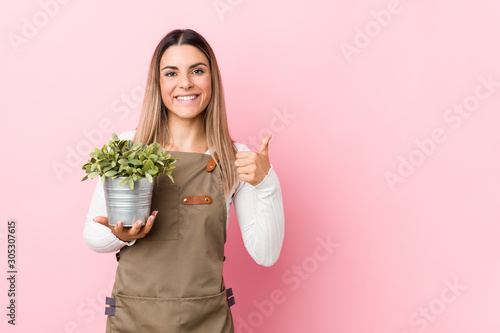Slika na platnu Young gardener woman holding a plant smiling and raising thumb up