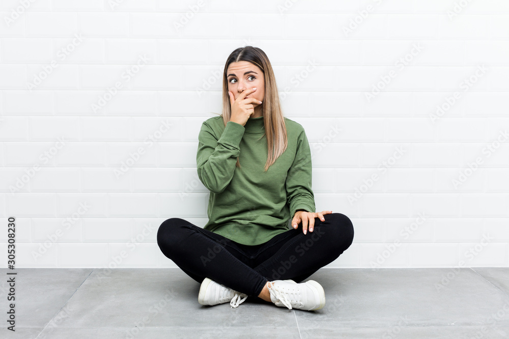 Young caucasian woman sitting on the floor thoughtful looking to a copy space covering mouth with hand. Young caucasian woman sitting on the floor thoughtful looking to a copy space covering mouth with hand.