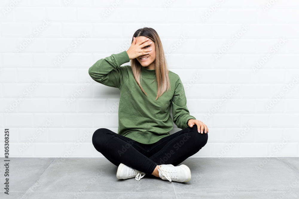 Young caucasian woman sitting on the floor blink at the camera through fingers, embarrassed covering face. Young caucasian woman sitting on the floor blink at the camera through fingers, embarrassed covering face.
