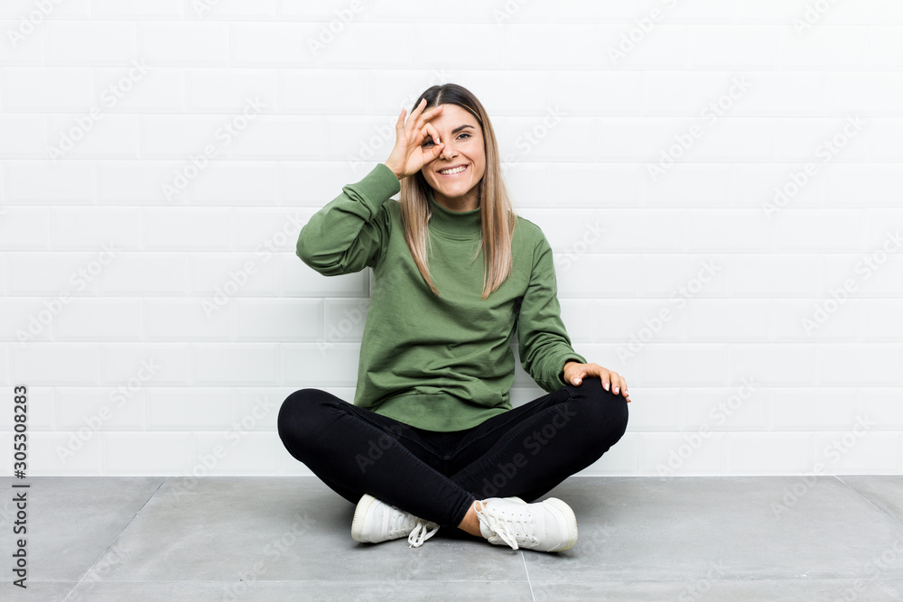 Young caucasian woman sitting on the floor excited keeping ok gesture on eye. Young caucasian woman sitting on the floor excited keeping ok gesture on eye.