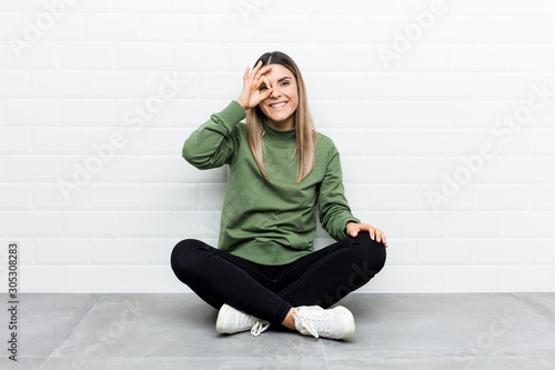 Young caucasian woman sitting on the floor excited keeping ok gesture on eye.