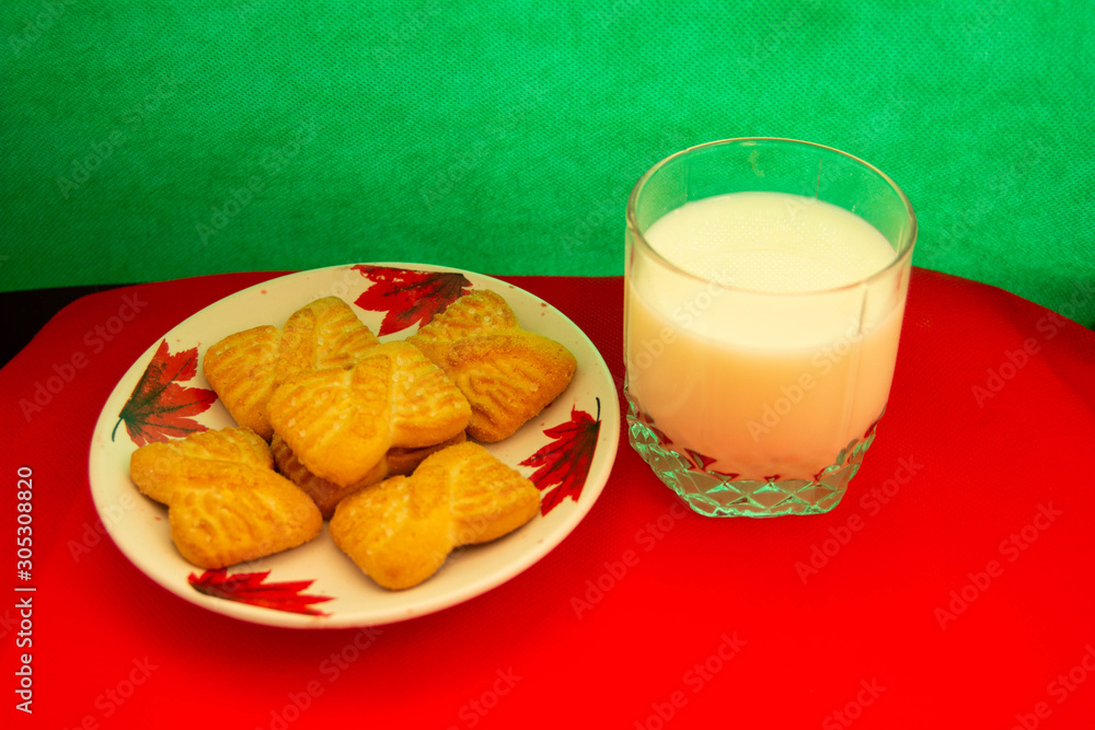 A glass of milk and cookies or shortcake biscuits on the plate with red and green background. National Cookie Day background. Christmas breakfast for Santa. American breakfast A glass of milk and cookies or shortcake biscuits on the plate with red and green background. National Cookie Day background. Christmas breakfast for Santa. American breakfast