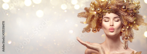 Christmas Woman with golden spruce tree wreath hairstyle on blurred pastel background. Beautiful Xmas model girl presenting hand, showing product, kiss. Hair style decorated, baubles. Christmas wishes