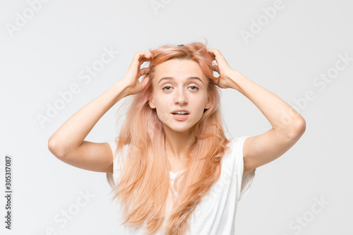 Close up studio portrait of young pretty blonde girl in white t-shirt. Woman raised hand to her hair and look at camera. An exclamation of surprise. Stunned puzzled female. Emotions concept.