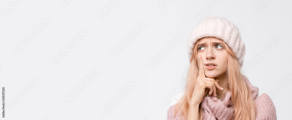 Studio portrait of attractive blonde girl in white hat and pink sweater. Worry female presses a finger to her cheek and thinks about problems in life. Emotions concept. Copy space. Studio portrait of attractive blonde girl in white hat and pink sweater. Worry female presses a finger to her cheek and thinks about problems in life. Emotions concept. Copy space.