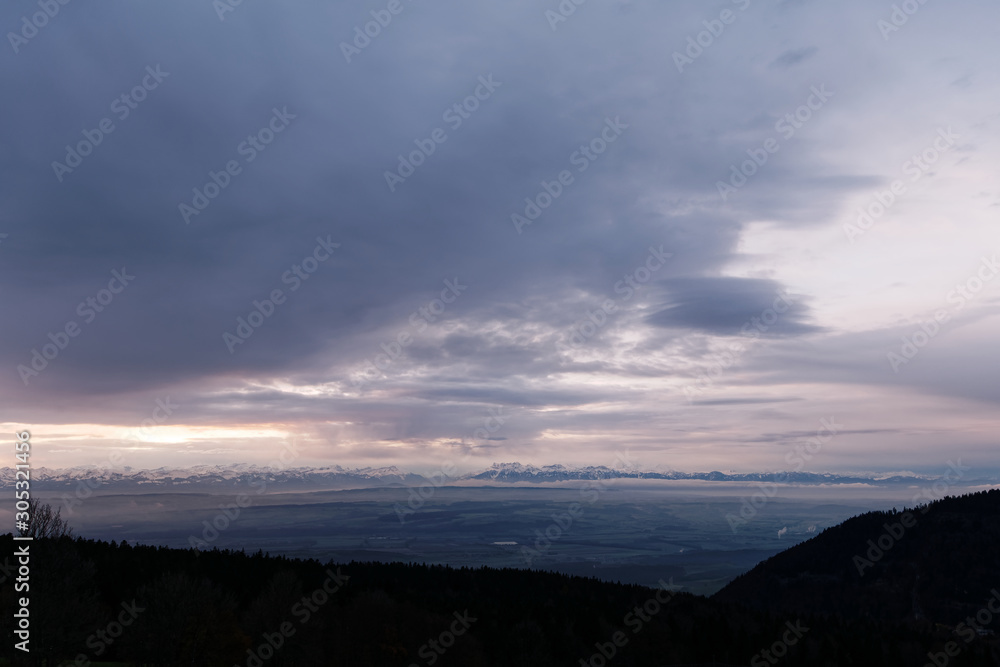 Fototapeta premium Winter landscape at sunrise. Silhouette of hills and trees in the foreground and snow capped mountain range of the European alps in the background.