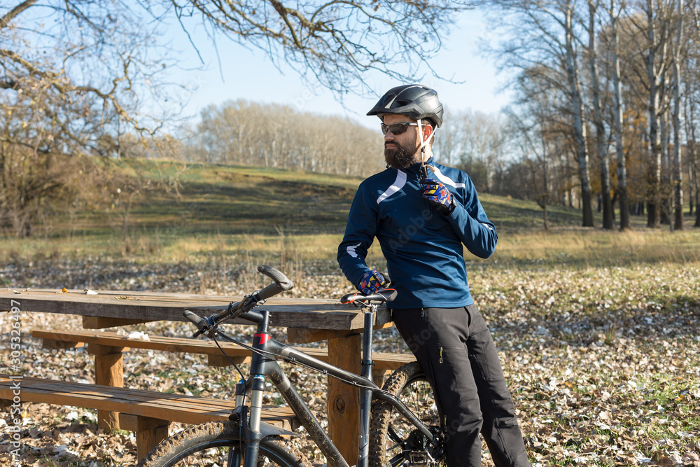 Cyclist in pants and fleece jacket on a modern carbon hardtail bike with an air suspension fork rides off-road. The guy is resting on a bench in the autumn park.