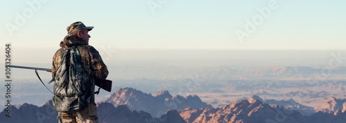 A hunter in camouflage and a gun in his hands in the early morning on top of a mountain stands looking for a target. Beautiful mountain landscape, the opening of the hunt.