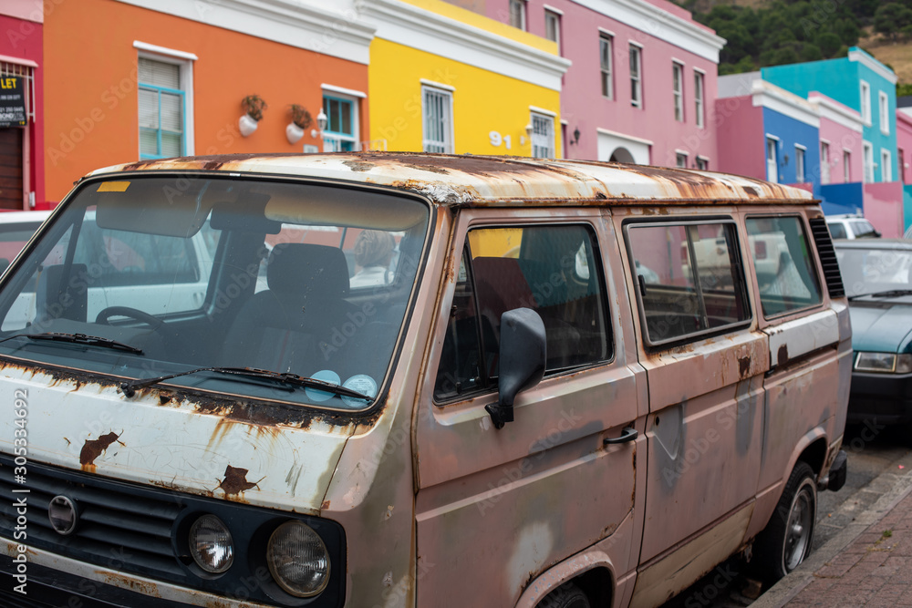 Old rusted car parked on street with colorful houses in background
