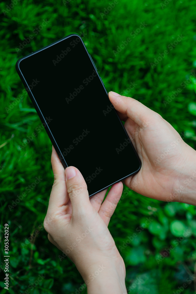 Close up woman hand holding and touching screen on the modern black smartphone mock up in vertical position with blank screen against blurred grass background. Clipping path inside. Close up woman hand holding and touching screen on the modern black smartphone mock up in vertical position with blank screen against blurred grass background. Clipping path inside.