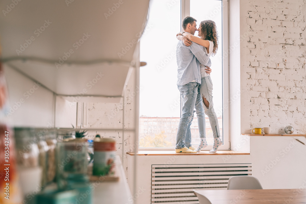 Young happy couple hugging on the windowsill in their apartment, sitting on the window in the sun Young happy couple hugging on the windowsill in their apartment, sitting on the window in the sun